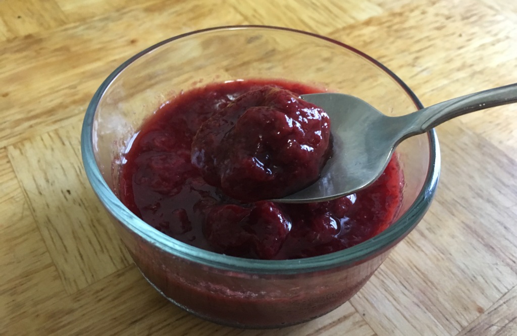 a stainless steel spoon displaying strawberry sauce over a small glass bowl of strawberry sauce on a wooden surface